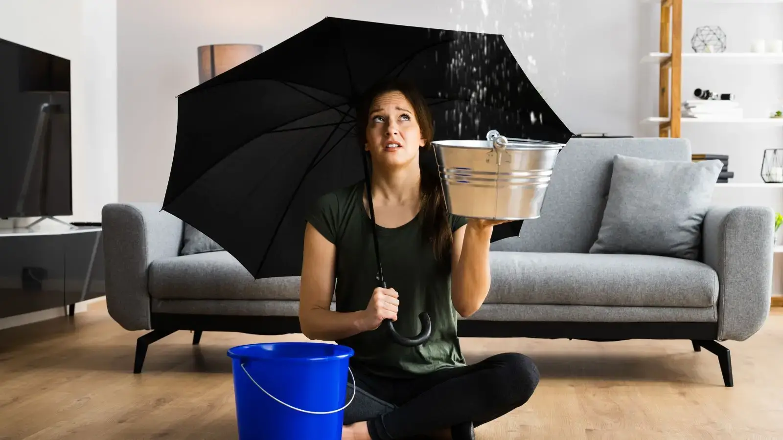 Woman holding umbrella and buckets to catch water leaks in living room, highlighting the importance of APS water sensor alarms