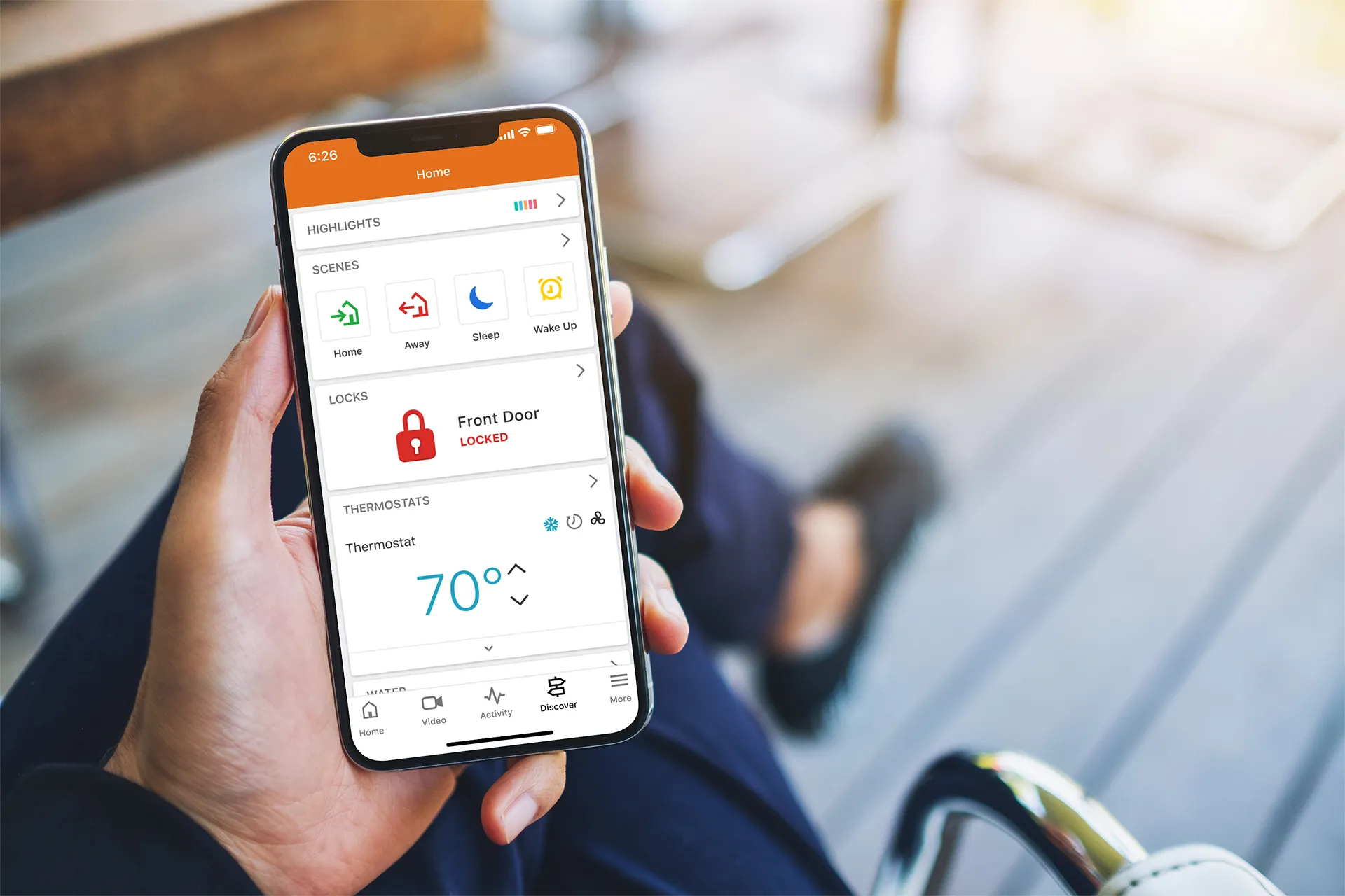 A person holding a smartphone displaying a home security app with smart home controls, including door locks, thermostat, and security scenes.