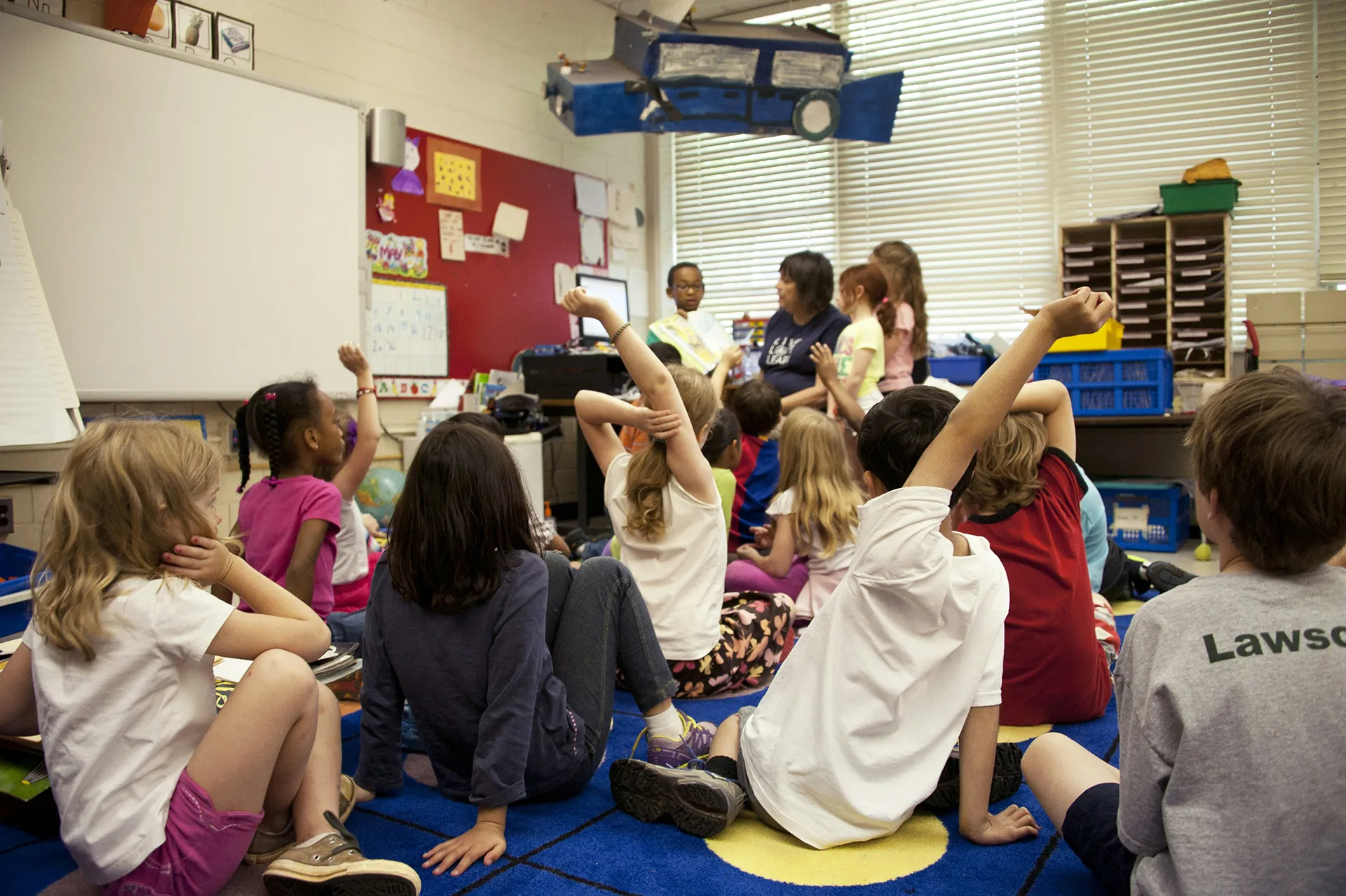 Elementary students raising their hands in a bright classroom as a teacher leads a lesson, symbolizing safe and engaging learning environments.